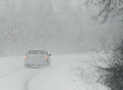 A car drives through heavy snow on a rural road.