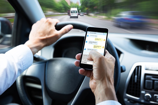 Driver holding the steering wheel with one hand while texting on a smartphone with the other, depicting distracted driving.